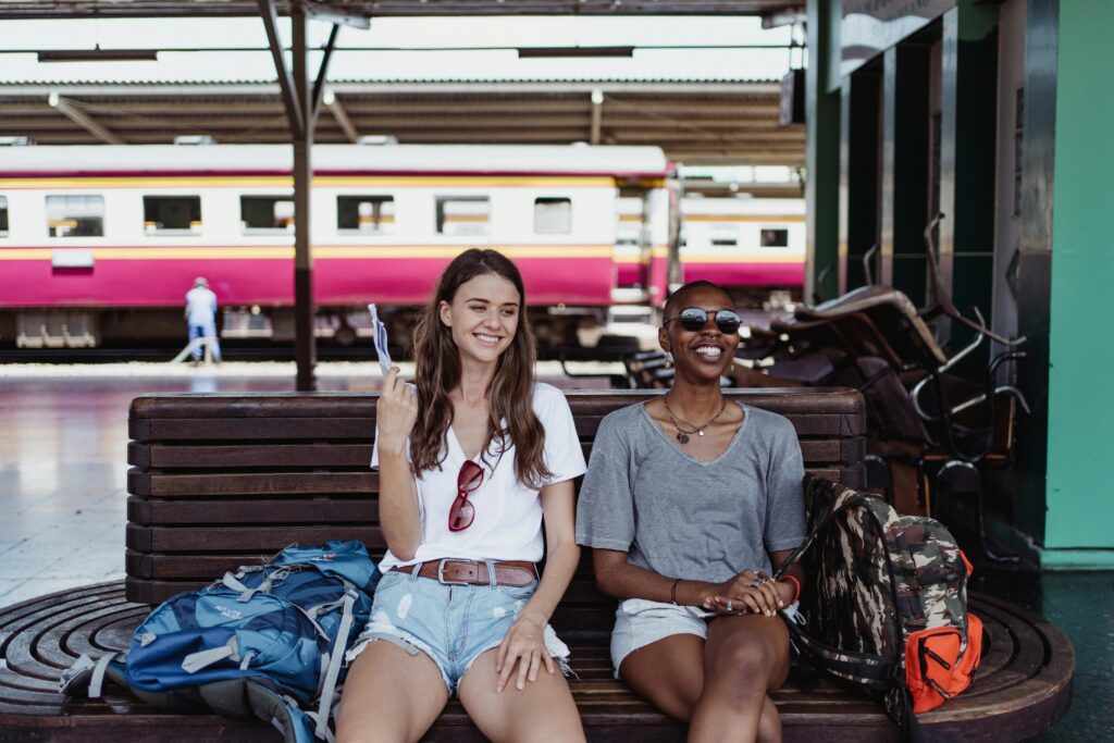 Two women backpackers smiling and waiting at a train station, sitting with their luggage.